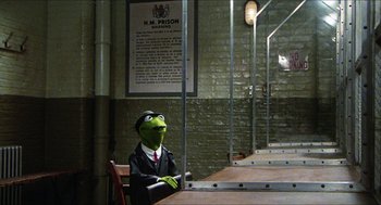 Movie still from “The Great Muppet Caper” (1981), directed by Jim Henson – A man dressed as kermit the frog sitting at a table; Wide shot, High angle