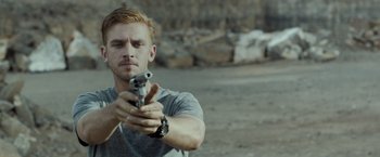 Movie still from “The Guest” (2014), directed by Adam Wingard – A man holding a gun while standing on a dirt field; Close Up shot, Over the shoulder angle