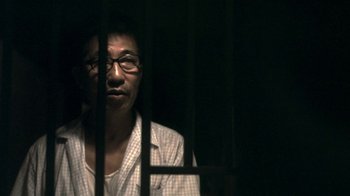 Movie still from “The Hand” (2004), directed by Kar-Wai Wong – A man with glasses is behind bars in the dark; Close Up shot, Over the shoulder angle