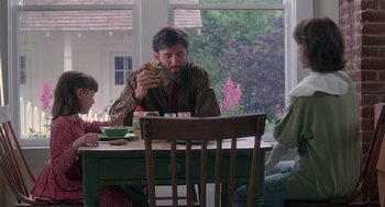 Movie still from “The Hand That Rocks the Cradle” (1992), directed by Curtis Hanson – A man sitting at a table eating a piece of food; Medium shot, Over the shoulder angle