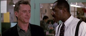 Movie still from “The Hard Way” (1991), directed by John Badham – Two men talking to each other in front of a group of people; Close Up shot, Over the shoulder angle