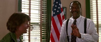 Movie still from “The Hard Way” (1991), directed by John Badham – A man standing in front of an american flag in a room; Medium shot, Over the shoulder angle