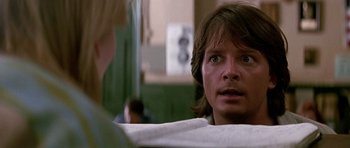 Movie still from “The Hard Way” (1991), directed by John Badham – A young man looking into the camera while sitting in a chair; Close Up shot, Over the shoulder angle