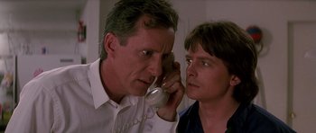 Movie still from “The Hard Way” (1991), directed by John Badham – A man on a phone with another man looking at him; Close Up shot, Low angle