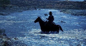 Movie still from “The Hired Hand” (1971), directed by Peter Fonda – A man riding on the back of a horse through water; Wide shot, High angle