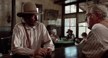 Movie still from “The Hired Hand” (1971), directed by Peter Fonda – A man sitting at a table in front of a group of people; Medium shot, Low angle