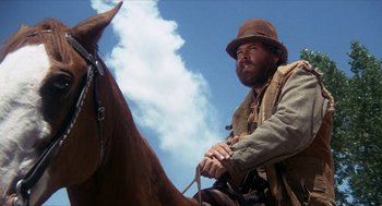 Movie still from “The Hired Hand” (1971), directed by Peter Fonda – A man sitting on top of a brown horse under a cloudy blue sky; Medium shot, Low angle