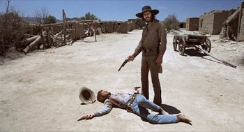 Movie still from “The Hired Hand” (1971), directed by Peter Fonda – A man in a cowboy hat is laying on the ground and another man is holding a gun pointed at him; Wide shot, Low angle