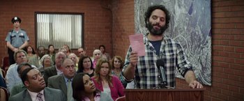 Movie still from “The House” (2017), directed by Andrew Jay Cohen – A man holding up a piece of paper in front of a crowd; Medium shot, Over the shoulder angle