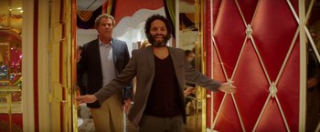 Movie still from “The House” (2017), directed by Andrew Jay Cohen – A man in a suit and tie standing next to another man in a suit and tie; Medium shot, Over the shoulder angle