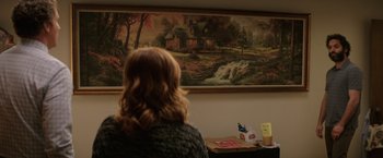 Movie still from “The House” (2017), directed by Andrew Jay Cohen – A woman sitting in front of a painting of a cabin; Wide shot, Over the shoulder angle