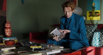Movie still from “The Human Voice” (2020), directed by Pedro Almodóvar – A woman sitting on a red couch looking at a magazine; Medium shot, High angle