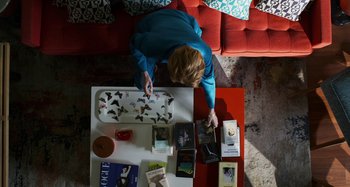 Movie still from “The Human Voice” (2020), directed by Pedro Almodóvar – A woman sitting at a coffee table looking at books; Wide shot, Overhead angle