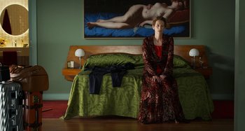 Movie still from “The Human Voice” (2020), directed by Pedro Almodóvar – A woman sitting on the edge of a bed in front of a painting; Wide shot, High angle