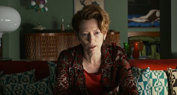 Movie still from “The Human Voice” (2020), directed by Pedro Almodóvar – An older woman sitting on a couch in a living room; Close Up shot, Over the shoulder angle