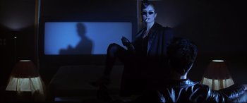 Movie still from “The Hunger” (1983), directed by Tony Scott – A woman with a cat face makeup sitting in front of a television; Medium shot, Over the shoulder angle