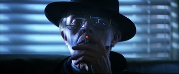 Movie still from “The Hunger” (1983), directed by Tony Scott – A man in a hat and glasses smoking a cigarette; Close Up shot, Low angle
