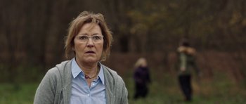 Movie still from “The Hunt” (2012), directed by Thomas Vinterberg – An older woman wearing glasses standing in a field; Close Up shot, Over the shoulder angle