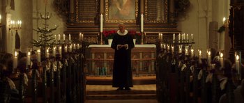 Movie still from “The Hunt” (2012), directed by Thomas Vinterberg – A man standing in front of candles in front of a painting; Wide shot, Low angle