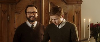Movie still from “The Hunt” (2012), directed by Thomas Vinterberg – A man standing next to a young man in front of a wooden wall; Medium shot, Over the shoulder angle