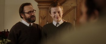 Movie still from “The Hunt” (2012), directed by Thomas Vinterberg – A young man in a sweater and tie standing next to an older man; Medium shot, Over the shoulder angle