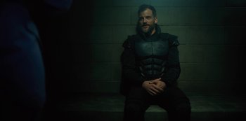 Movie still from “Altered Carbon” (2018), created by Laeta Kalogridis – A man in a black outfit sitting on a brick wall; Medium shot, Over the shoulder angle