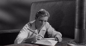 Movie still from “The Incredible Shrinking Man” (1957), directed by Jack Arnold – A young man is writing on a piece of paper while sitting on a chair; Medium shot, High angle