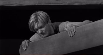 Movie still from “The Incredible Shrinking Man” (1957), directed by Jack Arnold – A man leaning on a wooden beam in a dark room; Medium shot, High angle