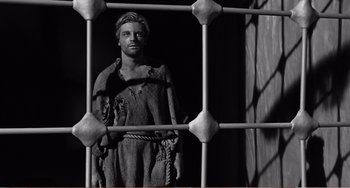 Movie still from “The Incredible Shrinking Man” (1957), directed by Jack Arnold – A statue of a man chained to a metal fence; Wide shot, Low angle