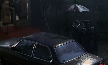 Movie still from “The Grilling” (1981), directed by Claude Miller – A person holding an umbrella over a car; Wide shot, High angle