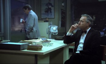 Movie still from “The Grilling” (1981), directed by Claude Miller – A man sitting at a table in front of a phone; Medium shot, Over the shoulder angle