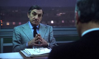 Movie still from “The Grilling” (1981), directed by Claude Miller – An older man sitting at a table in front of another man; Medium shot, Over the shoulder angle