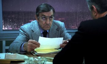 Movie still from “The Grilling” (1981), directed by Claude Miller – An older man sitting at a table with papers in front of him; Medium shot, Over the shoulder angle