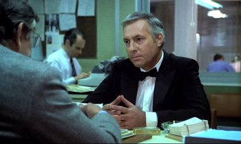 Movie still from “The Grilling” (1981), directed by Claude Miller – A man sitting at a table in front of another man; Medium shot, Over the shoulder angle