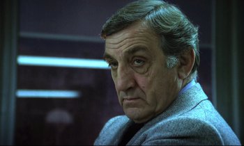 Movie still from “The Grilling” (1981), directed by Claude Miller – An older man in a gray jacket looks at the camera; Close Up shot, Low angle