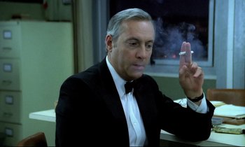 Movie still from “The Grilling” (1981), directed by Claude Miller – A man in a suit and tie holding a cigarette in his hand; Close Up shot, High angle
