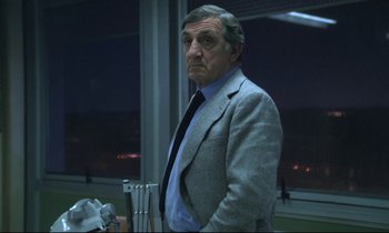 Movie still from “The Grilling” (1981), directed by Claude Miller – An older man wearing a suit and tie standing in front of a window at night; Medium shot, Low angle