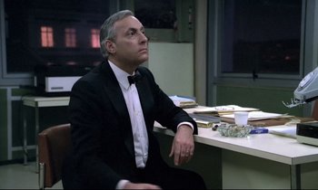 Movie still from “The Grilling” (1981), directed by Claude Miller – A man sitting at a table in front of a stack of papers; Medium shot, Low angle