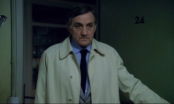 Movie still from “The Grilling” (1981), directed by Claude Miller – An older man in a trench coat and a tie; Close Up shot, Over the shoulder angle
