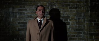 Movie still from “The Ipcress File” (1965), directed by Sidney J. Furie – A man in a suit and tie standing in front of a brick wall; Medium shot, Low angle