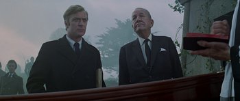 Movie still from “The Italian Job” (1969), directed by Peter Collinson – Two men in suits standing next to each other in front of trees; Medium shot, Low angle