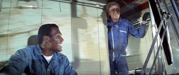 Movie still from “The Italian Job” (1969), directed by Peter Collinson – Two men in blue coveralls laughing in front of a window; Medium shot, Low angle