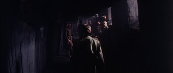 Movie still from “The Keep” (1983), directed by Michael Mann – A group of people standing around in a dark room; Wide shot, Over the shoulder angle