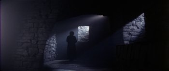 Movie still from “The Keep” (1983), directed by Michael Mann – A person standing in a dark room with a window; Wide shot, Low angle
