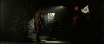 Movie still from “The Keep” (1983), directed by Michael Mann – A man standing next to a window in a dark room; Wide shot, Over the shoulder angle