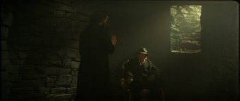Movie still from “The Keep” (1983), directed by Michael Mann – A man in a black coat and a man in a yellow hat; Wide shot, Over the shoulder angle