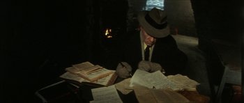 Movie still from “The Keep” (1983), directed by Michael Mann – A man in a hat and a suit sitting at a table with papers; Medium shot, High angle