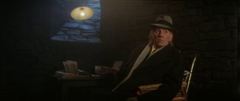 Movie still from “The Keep” (1983), directed by Michael Mann – A man in a hat sitting at a table in front of a lamp; Medium shot, Low angle