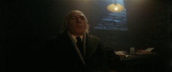 Movie still from “The Keep” (1983), directed by Michael Mann – An older man wearing a suit and tie in front of a light bulb; Close Up shot, Low angle