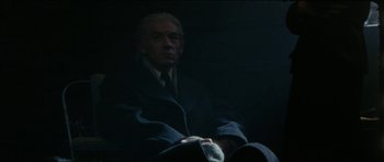 Movie still from “The Keep” (1983), directed by Michael Mann – A man in a suit and tie sitting in a dark room; Medium shot, Low angle